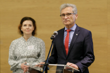 Close-up. Man in suit at lectern. Behind him, a woman. 