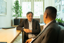 Two men are sitting in a room at a table. They are dressed in suits. One of them is facing the camera, the other with his back. They are talking.