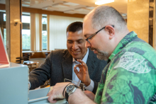 Two men in a library room. The one on the left dressed in a suit, the one on the right in a green shirt. They are looking at something together.