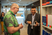 Two men in a library room. The one on the left is dressed in a suit, the one on the right in a green shirt. The one in the suit on the right has an open book in his hands. He smiles. 