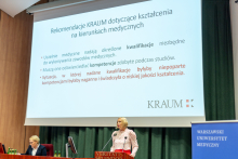 A woman, blonde, wearing a white blouse stands behind the lectern and speaks. Behind her a screen, on the screen - in black and red font - describes a recommendation.