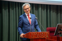 A man in a blue suit and red tie stands behind the lectern and smiles.