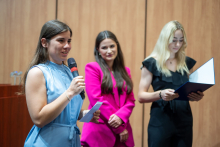 Three young girls standing in the middle of the room. The one on the left, wearing a blue dress, holds a microphone. The girl in the middle, dressed in a pink jacket is smiling. The girl on the right, dressed in black, is reading something from a document folder open in front of her.