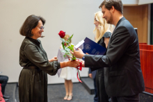 In the foreground, a young man presents a woman with a rose and a diploma. Behind them, a young girl, blonde, stands in the background. She is holding a briefcase.