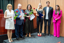 Group of people. They are standing in the hall. They are smiling at the camera. Some of them are holding flowers and diplomas.