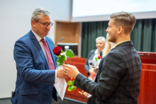 In the foreground, a young man presents another man with a rose and a diploma. Behind them, a young girl, blonde, stands in the background. 