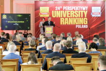 People sitting on chairs, with their backs to the camera. A man stands facing the camera, behind the lectern. To his left a screen. Behind him a wall with a red background and yellow lettering.