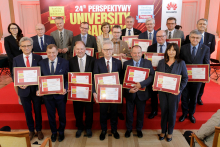 Group of people. Formally dressed. They stand and smile for a photograph. In their hands they hold framed diplomas.