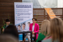 In the foreground is a woman with dark hair, wearing glasses and a pink jacket. She is sitting on a chair and talking. Behind her is a rollup with information about the Medical Center. In front of her, with her back to the camera, a group of listeners.