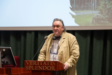 A man in a beige jacket at the lectern.