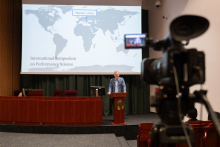 Lecture hall. At the lectern a man in a blue jacket. Behind him a screen showing a map of the world.