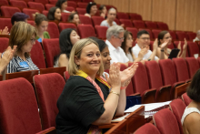 Listeners sit in chairs, applaud. In the foreground is a smiling woman.