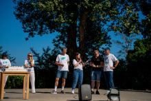 Sześć osób stoi na boisku do siatkówki jak na scenie. Pięć z nich ma koszulki z napisem "2023 Chłapowo". Dziewczyna z długimi czarnymi włosami mówi do mikrofonu.