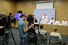 Facing the camera a group of people - they are sitting at a conference table, and one of them is standing. He holds a microphone in his hand and speaks into it. With their backs to the camera, a group of journalists stand behind cameras and sit in chairs.