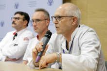 Three men sitting at a table. All dressed in white aprons and wearing glasses. One of them has a microphone in his hand and is speaking into it, with his other hand pointing to something on the table.