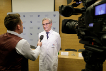 A man in a white apron and glasses stands and says something into a microphone. In front of him stands another man, who holds this microphone in his hand. On the side, to the right - a TV camera.