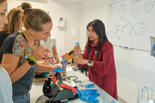 On the table lies expert airway equipment. On one side, a woman in a red dress demonstrates how to operate it. On the other side of the table, several young women - course participants - are watching.