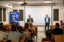 Lecture hall. From the back, the heads of the audience are visible. At the end of the room, facing the audience two men in suits. Behind them a screen.