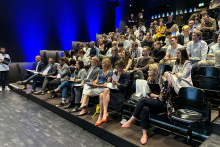 A group of people sitting on chairs in a large hall. They are looking in one direction.