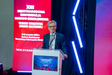 A smartly dressed man stands behind a lectern and speaks into a microphone. In the background behind him is a screen - a telescreen with the name of the conference