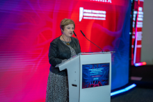 A smartly dressed woman stands behind a lectern and speaks into a microphone. In the background behind her is a screen - a telescreen with the name of the conference