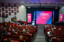 Lecture hall. People sitting on chairs, a man stands in front of them and speaks behind the lectern. Behind him a huge screen with the name of the conference