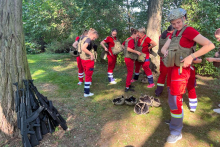 Kilka kobiet stoi na polania. Mają na sobie czerwone stroje ratowników. Zakładają kamizelki kuloodporne. Niektóre mają kaski. 