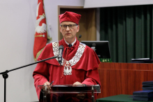 A man in a red toga and red biretta at the lectern, speaks into the microphone.