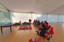 A large room, in a row on chairs sit women. in front of them a blanket, on which lies a dummy, and a woman performs ventilation.