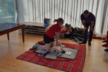 A blanket is spread on the floor. On the blanket, a young woman is practicing chest compressions on a mannequin. An older man is watching.