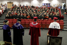 Aula. Na scenie tyłem cztery osoby w togach, dalej stoi publiczność, również w togach i biretach.