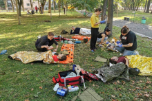 Na łące leżą trzy osoby odgrywające rannych. Są przykryte foliami termoizolującymi. Przy nich osoby udzielające pomocy.