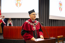 Mężczyzna w średnim wieku w czerwono-czarnej todze, okularach i czapce akademickiej mówi do mikrofonu. 