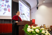 A young woman wearing glasses and dressed in a black and red toga speaks into a microphone. Behind her a multimedia screen with a colour slide. In front of the lectern a basket of white roses.