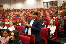 Młody mężczyzna stoi i coś mówi. Obok niego, przed nim, za nim, w rzędach - siedzą młodzi ludzie, ubrani elegancko.