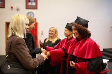 Several people, two women in the foreground - a woman with light hair receives something from a woman with dark hair, dressed in an academic toga.