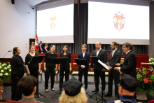 A group of people dressed in black stand at the centre of the room, singing. Behind them is a multimedia screen with the department's logo displayed in maroon.