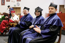 Three people dressed in navy blue and black academic togas sit in chairs, behind them a middle-aged man in a red and white toga sits in an armchair.