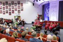 View of the auditorium, with people of different ages in the seats, in the foreground a person standing behind the lectern, and decoration in the background.