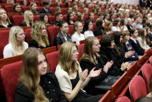 Młode osoby ubrane w stroje galowe siedzą na czerwonych krzesłach i biją brawo.