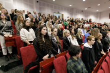 A group of people, dressed variously. They stand upright in rows with chairs