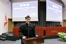 A middle-aged man in a black academic toga stands behind a lectern. Behind him a multimedia screen with a colour slide projected.