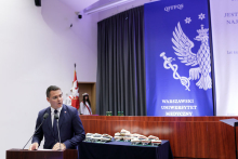 A middle-aged man, wearing a suit and tie, stands behind the rostrum, his head turned to the left. Behind him, an inaugural decoration in white and blue with an eagle.