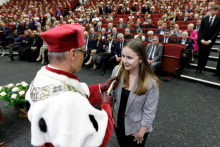 W tle wiele osób siedzących na czerwonych fotelach. Na pierwszym planie młoda dziewczyna ubrana elegancko, uśmiecha się, tyłem do aparatu starszy mężczyzna, który berłem rektorskim dotyka jej ramienia.