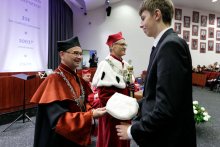 A tall young man dressed in a suit receives his cap - the symbol of matriculation - from a middle-aged man dressed in a black and orange dean's toga.