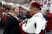 In the foreground, on the left a young girl with fair hair dressed elegantly, on the right a middle-aged man in a red and white toga touches her right shoulder with a rector's sceptre. There are many people in the background.