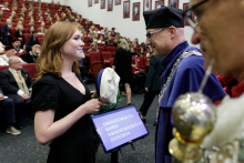 On the left, a young girl in a black dress receives a cap - a symbol of matriculation - from a middle-aged man in a black and blue toga. Many people in the background.