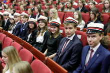 A group of young people dressed elegantly, sit in red chairs. They are smiling at the camera. On their heads they are wearing white caps with a red brim and a black visor.