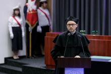 A young man in a black toga, wearing glasses, stands behind the rostrum. Behind him is a banner post, with a grey curtain in the background.