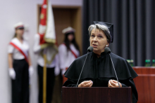 A middle-aged woman, dressed in a black academic toga, speaks into a microphone. Behind her, a banner post, a grey curtain.